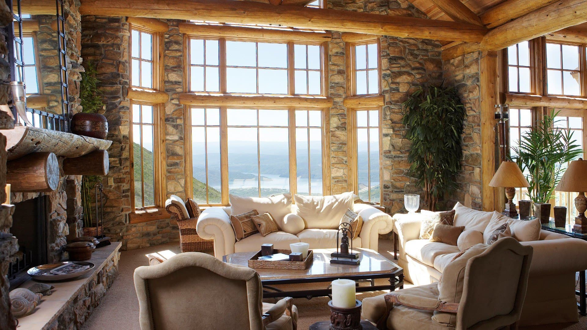 Rustic living room with stone walls, log beams, and panoramic windows overlooking a scenic lake.