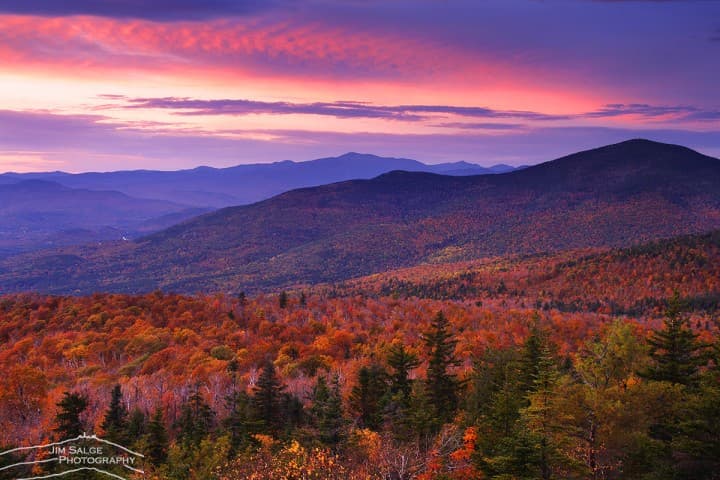 Vibrant pink sunset over rolling mountains with colorful autumn foliage and scattered evergreen trees.