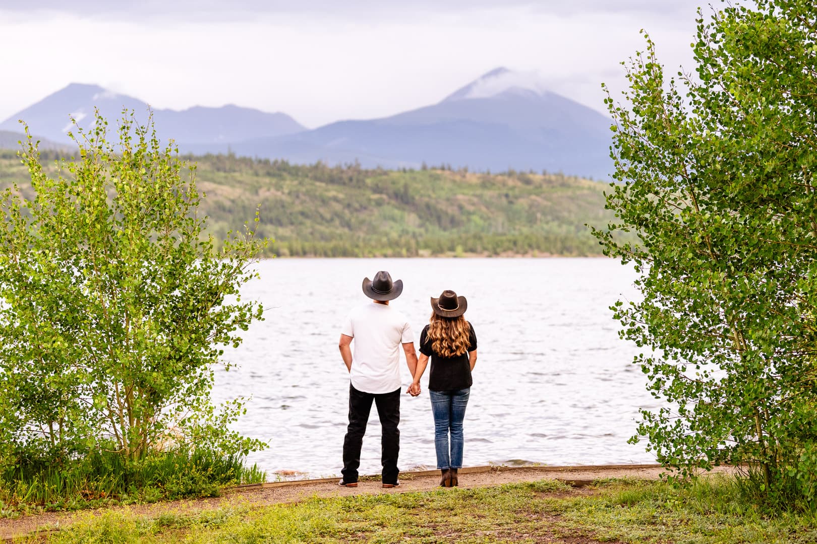 Couple in cowboy hats holding hands, looking at a mountain lake between green trees.