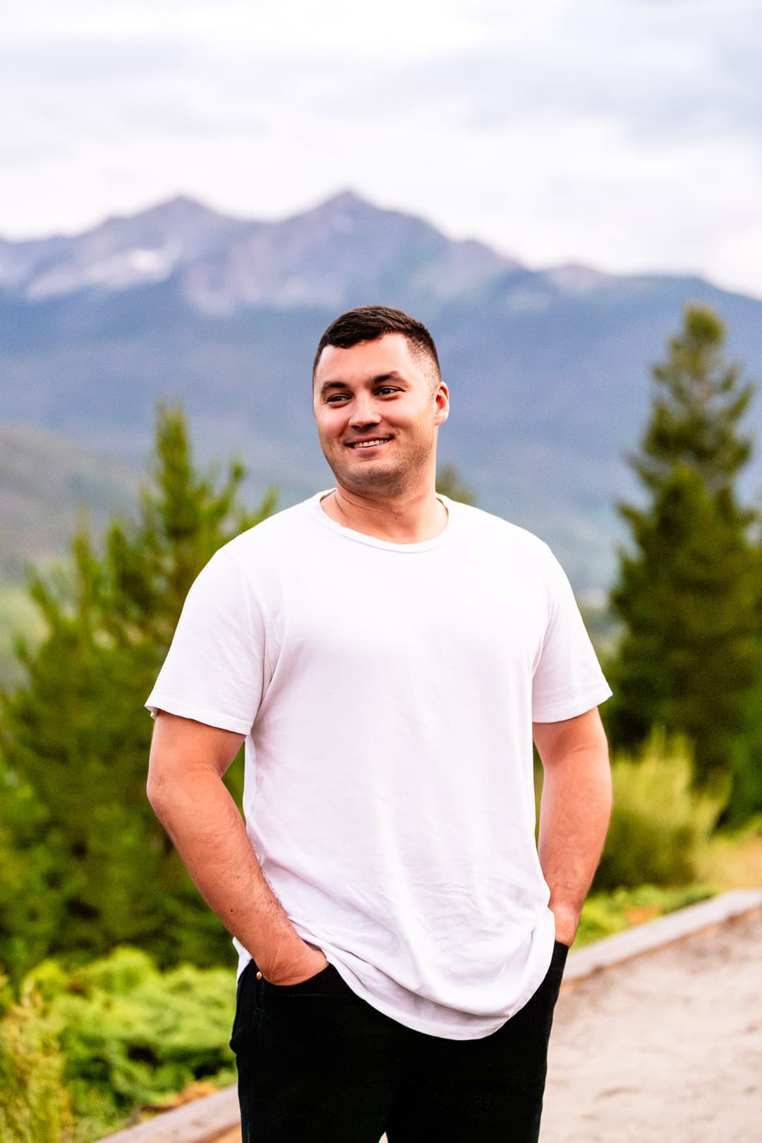 Smiling man in a white t-shirt stands outdoors with a scenic mountain range background.