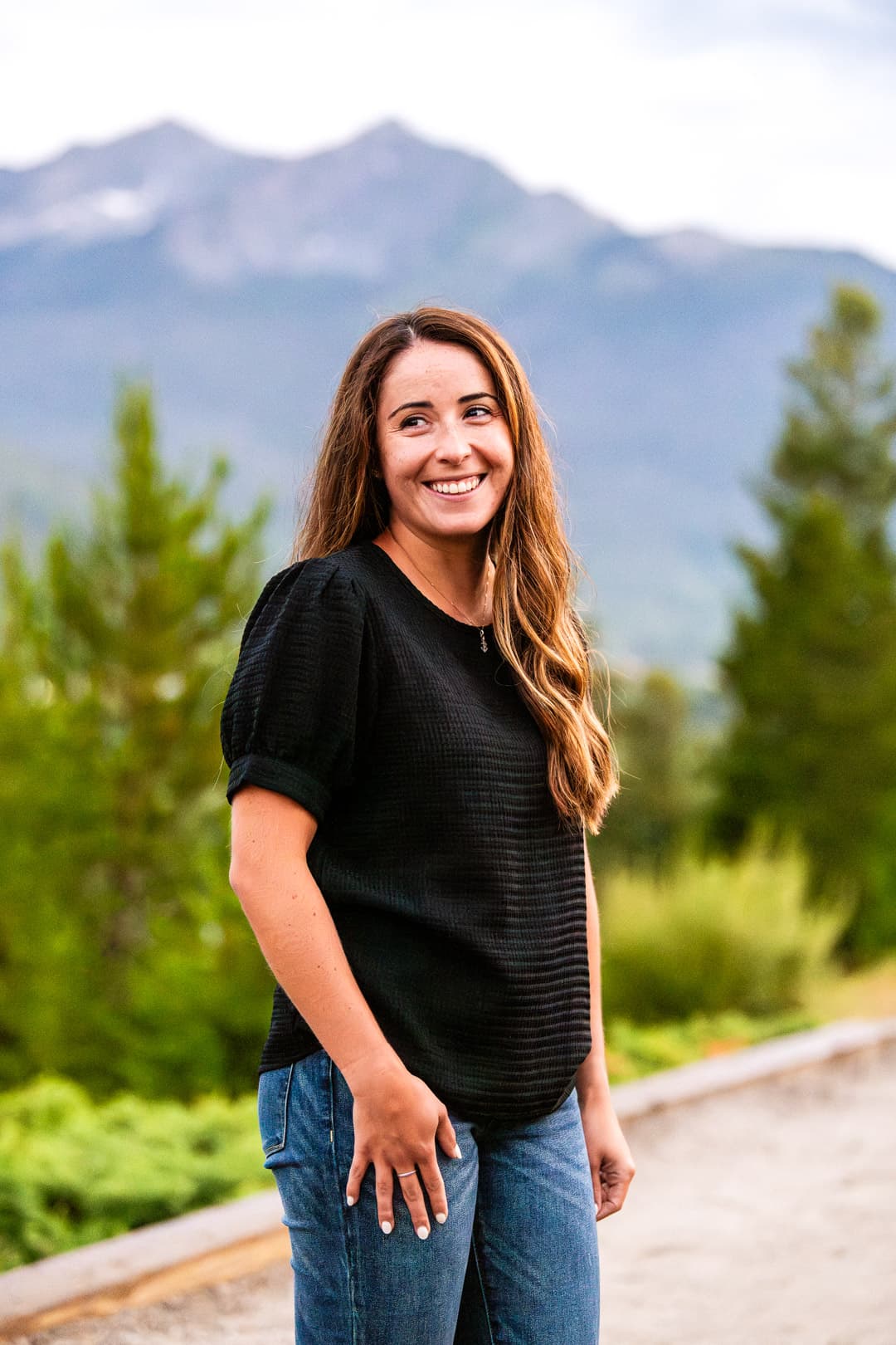 Smiling woman with long brown hair in a black top, with mountains in the background.