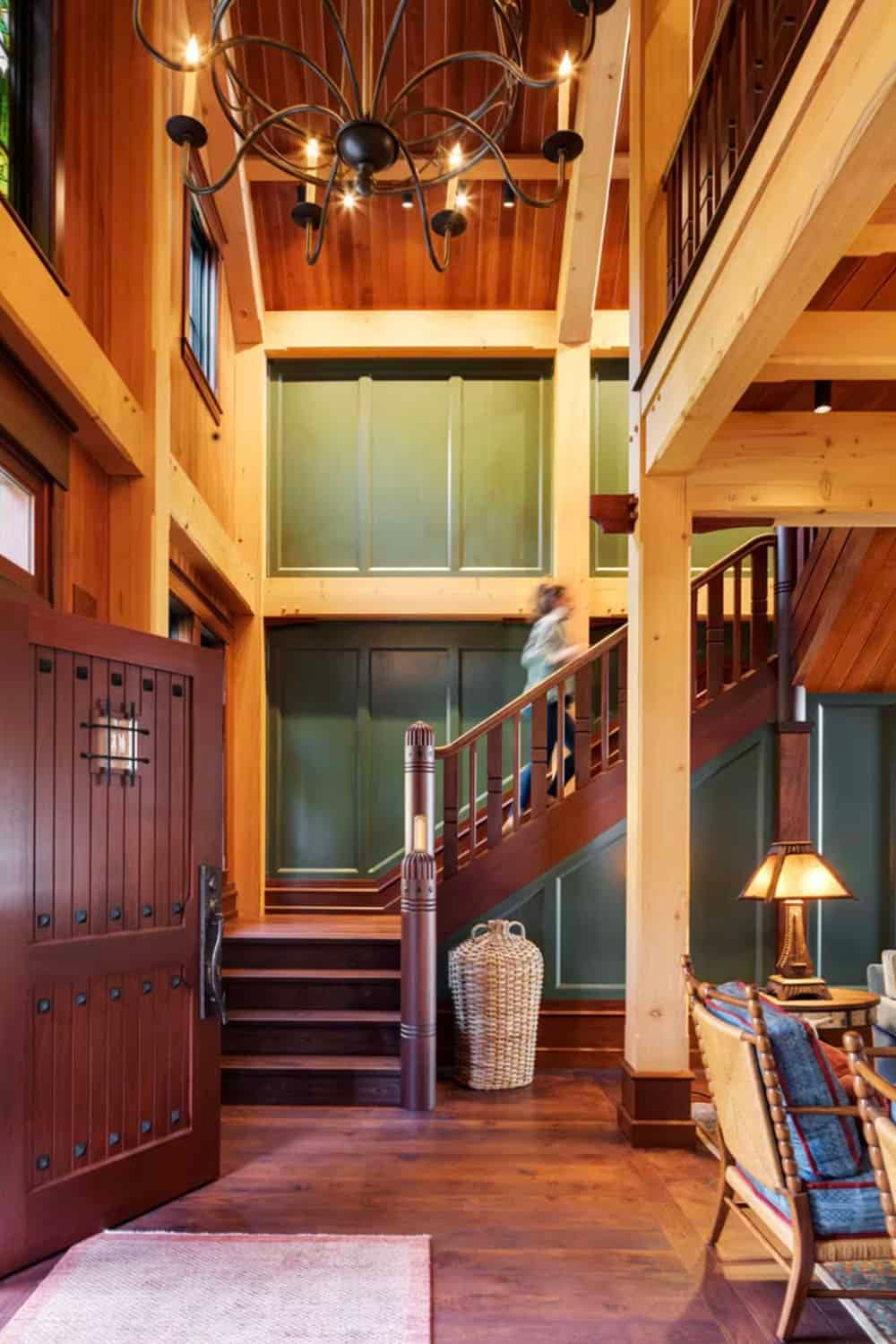 Rustic timber-frame foyer with a wooden staircase, green paneled walls, and an iron chandelier.
