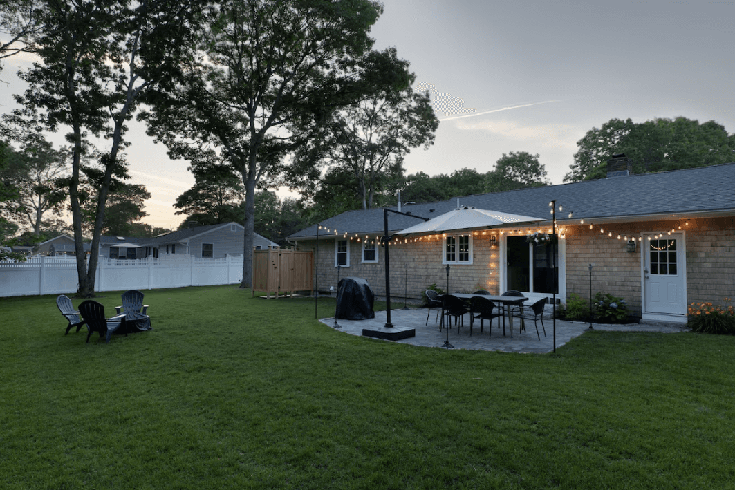 Backyard patio with string lights, dining set, and Adirondack chairs on a lawn at dusk.