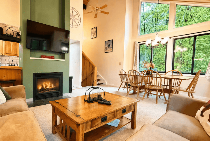 Cozy living room with green fireplace wall, wooden furniture, and dining area with large windows.