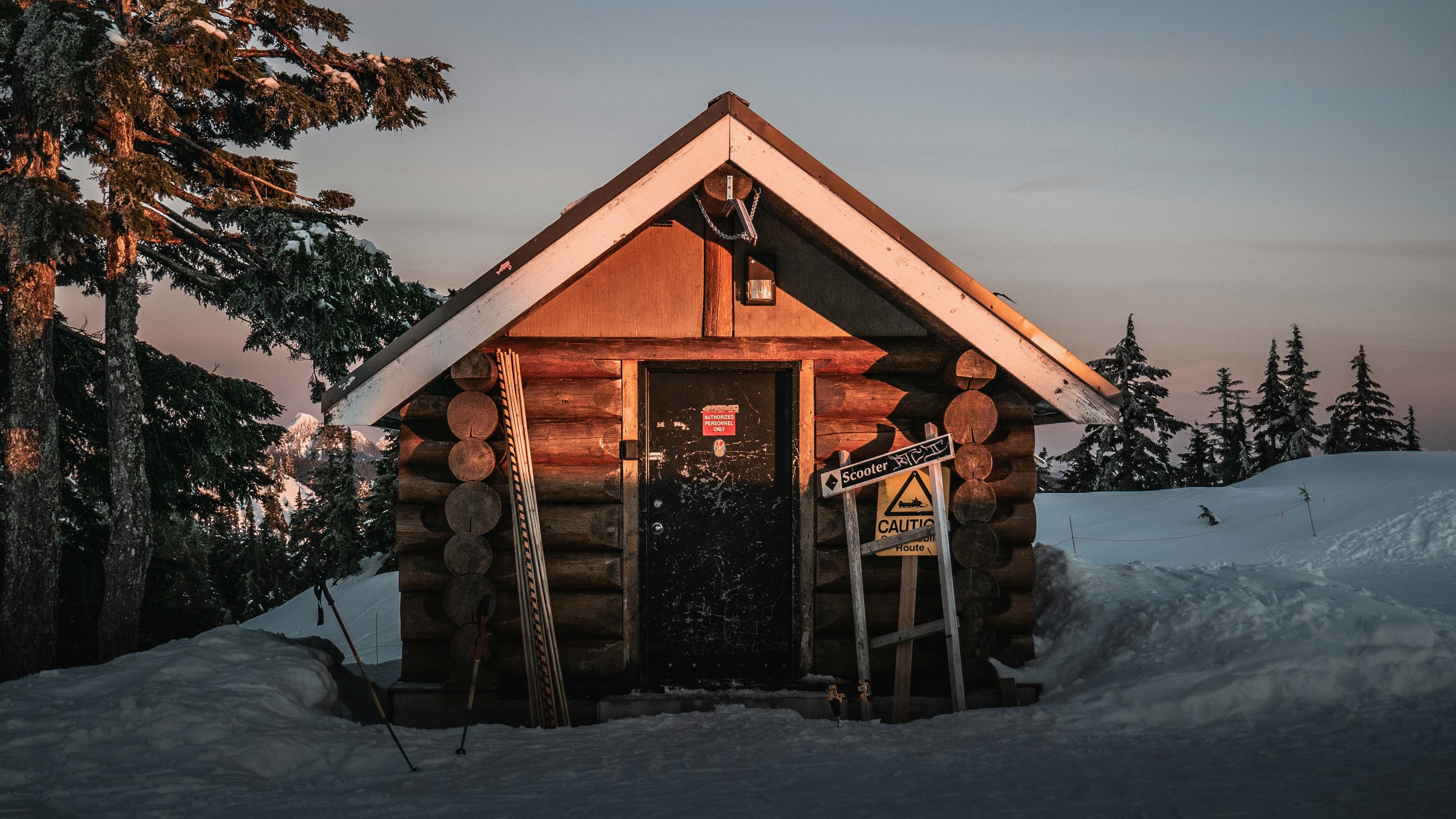Rustic log cabin nestled in deep snow at sunset, surrounded by pine trees and mountains.